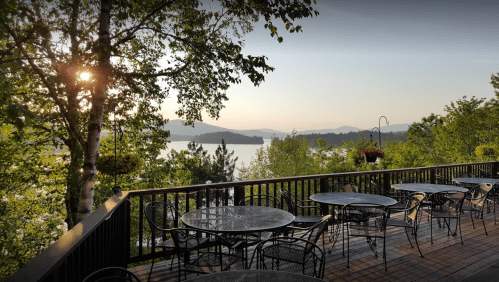 A scenic view from a deck with empty tables, overlooking a lake and mountains at sunset.
