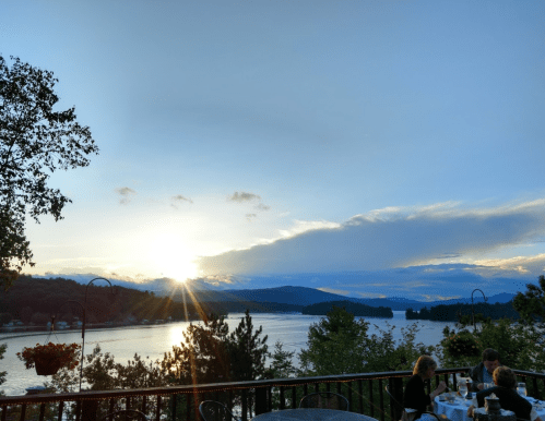 Sunset over a tranquil lake, with silhouettes of trees and people dining on a deck in the foreground.
