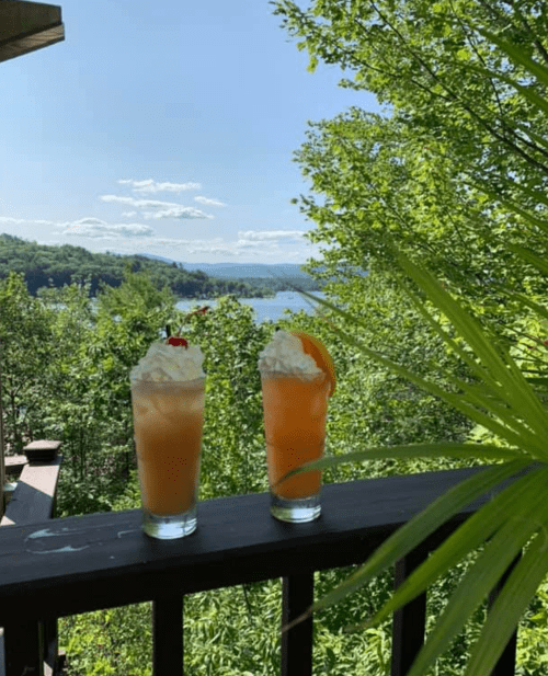 Two colorful cocktails with whipped cream and garnishes sit on a wooden railing, overlooking a scenic lake and lush greenery.