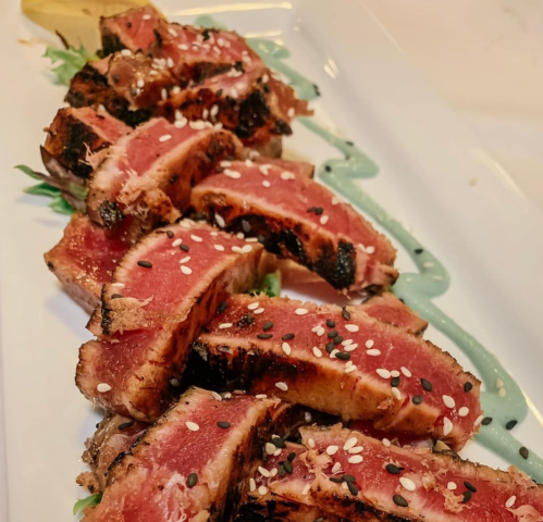 Sliced seared tuna arranged on a plate, garnished with green sauce and sprinkled with sesame seeds.