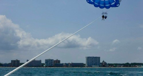 Two people parasailing over the ocean with a city skyline in the background under a partly cloudy sky.