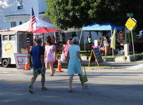 Two people walk on a street near food stalls and tents at a community event, with an American flag in the background.