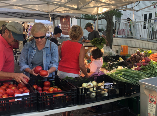 A bustling farmers market with people selecting fresh produce, including tomatoes, greens, and root vegetables.