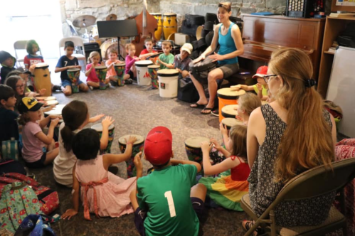 A group of children sitting in a circle, playing drums, with an instructor leading the session in a music room.