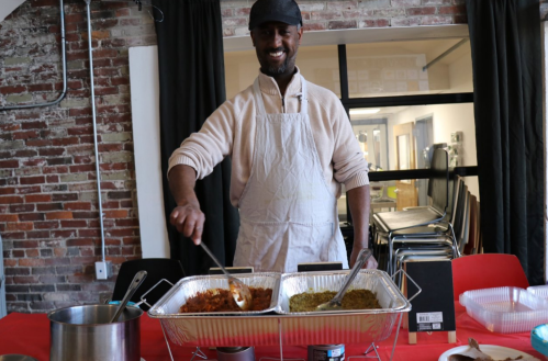 A smiling man in an apron stirs spices in large trays at a table, with a brick wall and chairs in the background.