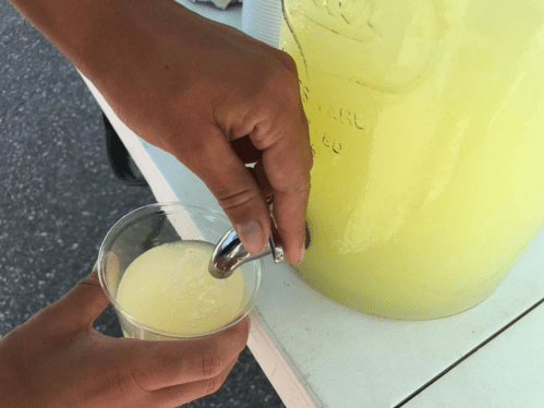 A hand pours a light yellow beverage from a large container into a clear cup.