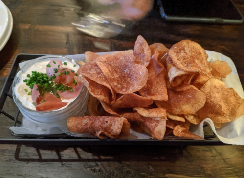A basket of crispy potato chips served with a small dish of creamy dip topped with chives and diced tomatoes.