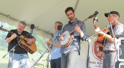 Four musicians perform on stage under a tent, playing guitar and banjo at a music festival.