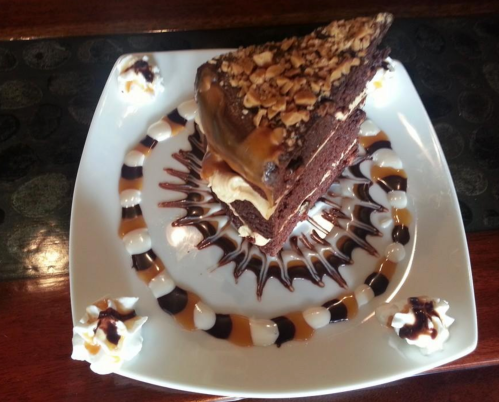A slice of chocolate cake topped with nuts and caramel, surrounded by decorative chocolate and whipped cream.