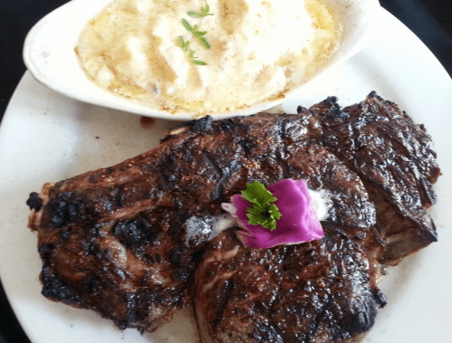 A plate featuring grilled steak with a garnish of purple flower, alongside a dish of creamy baked potatoes.