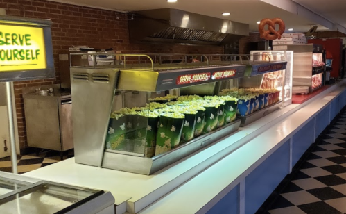 A concession stand featuring large popcorn containers, a pretzel display, and a "Serve Yourself" sign in a retro setting.