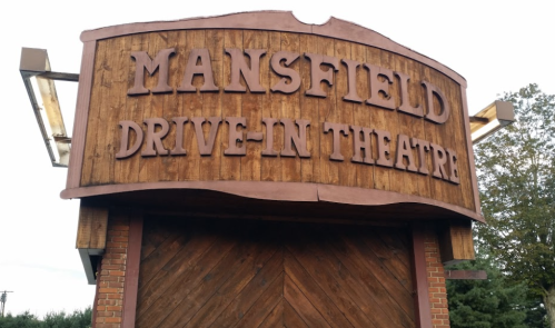 Sign for Mansfield Drive-In Theatre, featuring a wooden design and bold lettering against a cloudy sky.