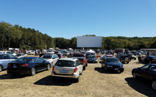 A busy outdoor drive-in theater parking lot filled with cars under a clear blue sky. A large blank screen is visible.