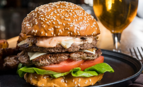 A stacked burger with sesame seed bun, lettuce, tomato, cheese, and sauce, served with a glass of beer in the background.