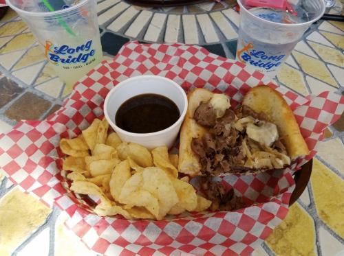 A basket with a sandwich filled with meat and cheese, served with chips and a cup of dipping sauce, on a tiled table.