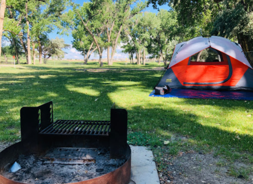 A campsite with a fire pit in the foreground and a red and gray tent set up on green grass under a blue sky.