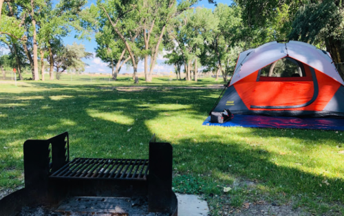 A campsite with a grill in the foreground and a red and gray tent set up on green grass under trees.