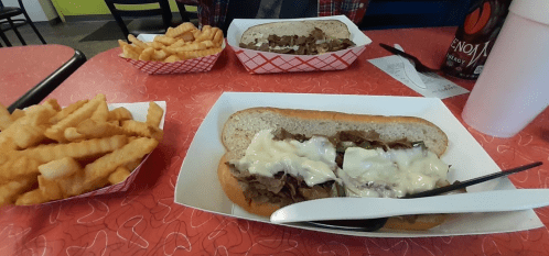 A table with a sandwich filled with meat and cheese, crinkle-cut fries, and a drink in the background.