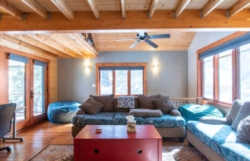 Cozy living room with wooden beams, a large sofa, bean bags, and a red coffee table, featuring natural light from windows.