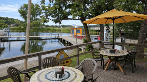 A lakeside view featuring a wooden deck with tables, an umbrella, and a dock in the background.