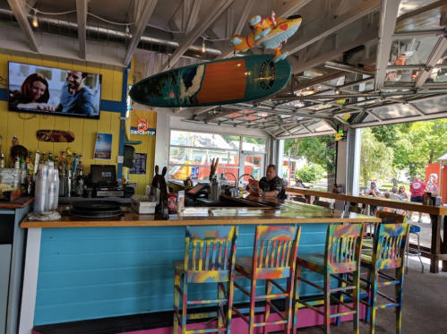 A colorful bar with a surfboard hanging above, featuring stools and a TV showing a couple enjoying a drink.