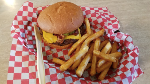 A cheeseburger with bacon on a bun, served with golden French fries in a red and white checkered basket.