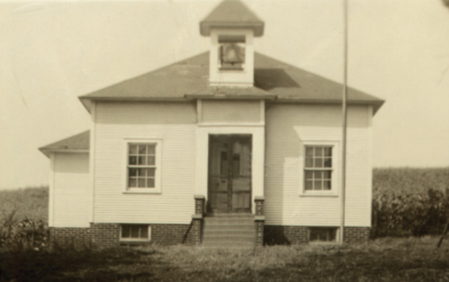 A vintage one-room schoolhouse with a bell tower, surrounded by fields and featuring a set of stairs at the entrance.