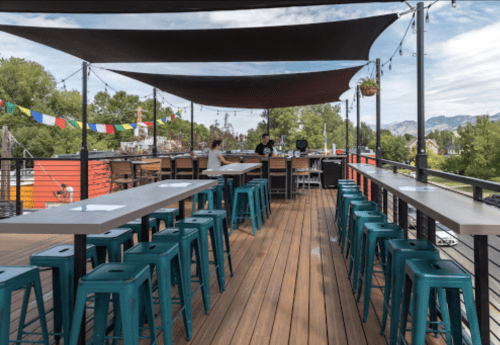 A rooftop bar with wooden flooring, teal stools, and a view of mountains, featuring colorful flags and shaded seating.