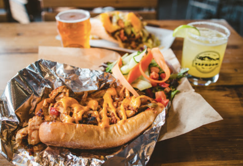 A hot dog topped with cheese sauce, served with a side salad and drinks on a wooden table.