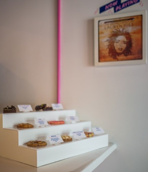 A display of assorted cookies on a white stand, with labels, next to a framed album cover on the wall.