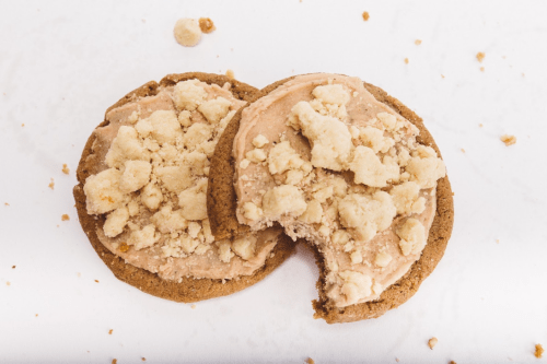 Two cookies with a creamy topping and crumbled pieces on top, one partially bitten, on a white surface.