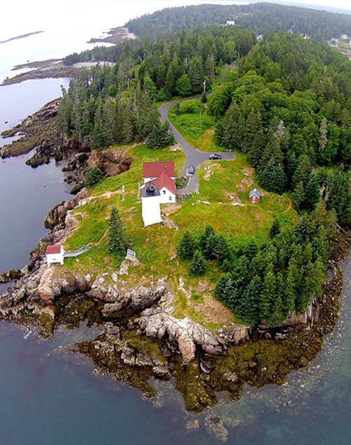 Aerial view of a lighthouse on a rocky island surrounded by trees and water, with a winding road and small buildings.