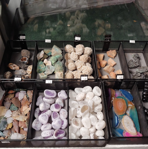 A display of various gemstones and minerals in black mesh trays, arranged by type and color.