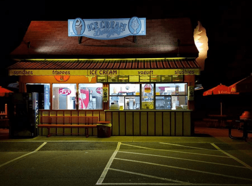 A brightly lit ice cream stand at night, featuring a menu of treats and an "OPEN" sign. Outdoor seating is visible.