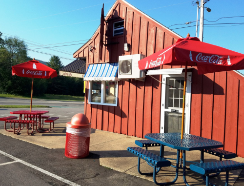 A red wooden building with blue and red outdoor seating, featuring Coca-Cola umbrellas and a trash can nearby.