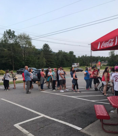 A long line of people waiting outside a building, with cars parked nearby and trees in the background.