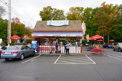 A colorful ice cream shop with outdoor seating, surrounded by trees in autumn colors, and customers at the counter.