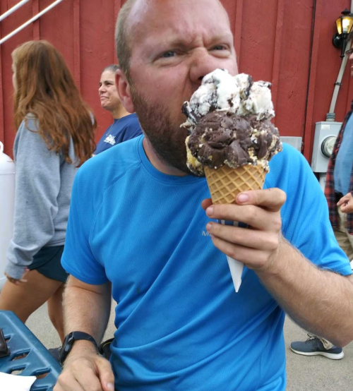 A man in a blue shirt playfully pretends to bite a large ice cream cone with multiple scoops.