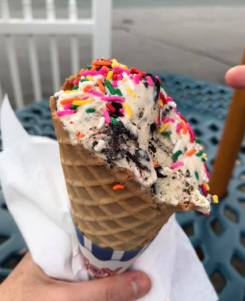 A hand holding a cone filled with ice cream topped with colorful sprinkles and cookie pieces.