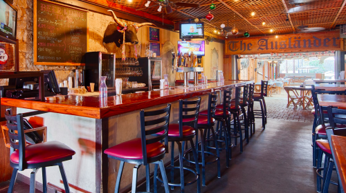 A rustic bar interior with wooden counters, high stools, and a warm atmosphere, featuring TVs and a chalkboard menu.