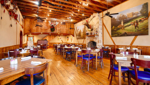 Cozy restaurant interior with wooden beams, tables set for dining, and rustic decor featuring paintings and wall hangings.