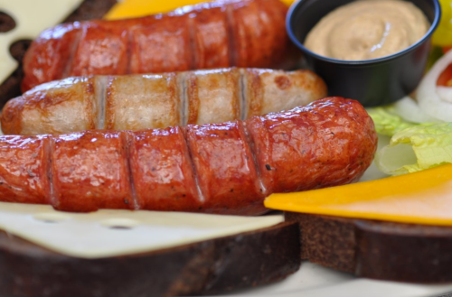 Three grilled sausages on a plate with cheese, lettuce, and a small bowl of mustard.