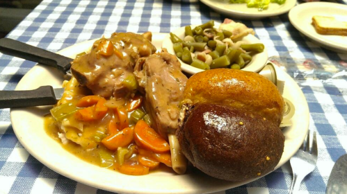 A plate of savory meat with gravy, carrots, green beans, and two types of bread on a checkered tablecloth.