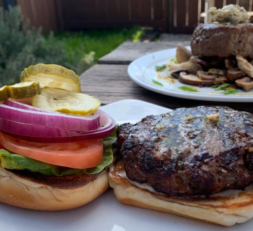 A close-up of a juicy burger with lettuce, tomato, onion, and pickles on a toasted bun, with a side dish in the background.