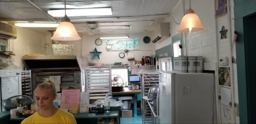 A cozy bakery interior with a counter, ovens, and a staff member in a yellow shirt, illuminated by pendant lights.