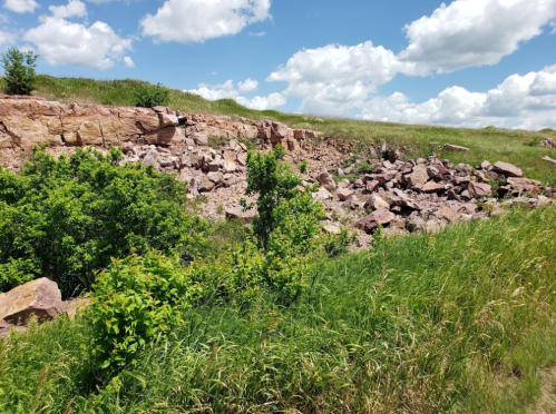 A rocky hillside with green grass and shrubs under a blue sky with fluffy white clouds.