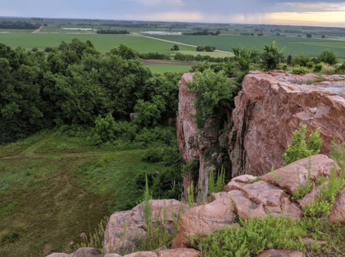 A rocky cliff edge overlooking lush green fields and trees under a cloudy sky.