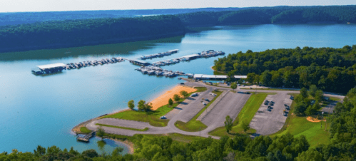 Aerial view of a marina on a lake, surrounded by green trees and a parking area, with boats docked in the water.