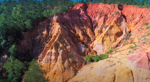 Vibrant red and orange rock formations with green trees on a steep hillside under a clear blue sky.
