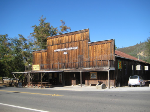 Historic wooden building with a sign for "Gold Trail Grange" and a café, set against a clear blue sky.